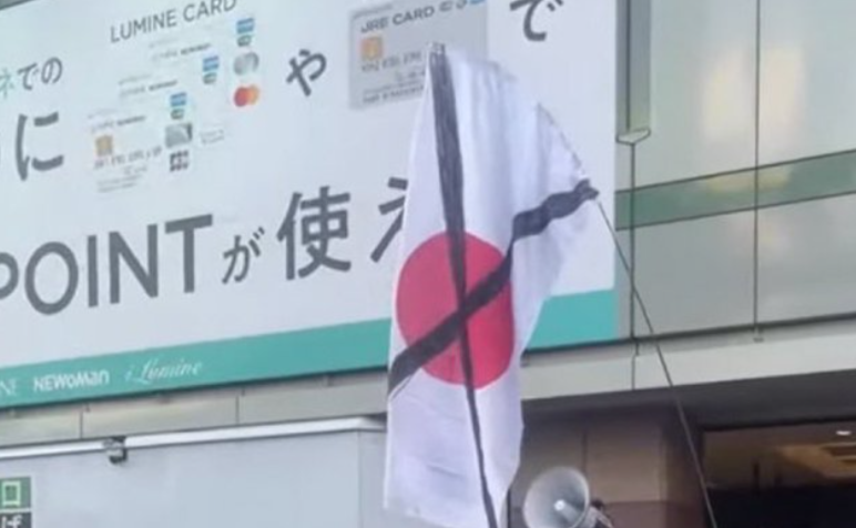 The image shows a white flag with a red circle in the center, resembling the Japanese flag, but it has two black diagonal lines crossing over the circle, one from the top left to bottom right and another from the top right to bottom left. The flag is mounted on a pole and is displayed in front of a building with a large sign featuring Japanese text and various logos, including credit card symbols. Part of the building’s exterior and a large speaker mounted below the sign are also visible.