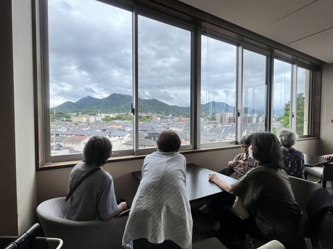 窓際で景色を眺める入居者たち。広い窓から山並みと町並みが見える共用スペース