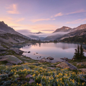 A sweeping sunrise vista unfolds across sapphire‑tinted valleys covered in soft morning dew, with alpine wildflowers glowing in fresh color. A crystal‑clear lake reflects the pastel sky as granite mountain peaks catch the first warm sunlight. Gentle mist drifts above the water between scattered evergreens and weathered shoreline boulders. The scene feels calm and expansive, with soft lavender and peach clouds stretching across the early morning horizon.