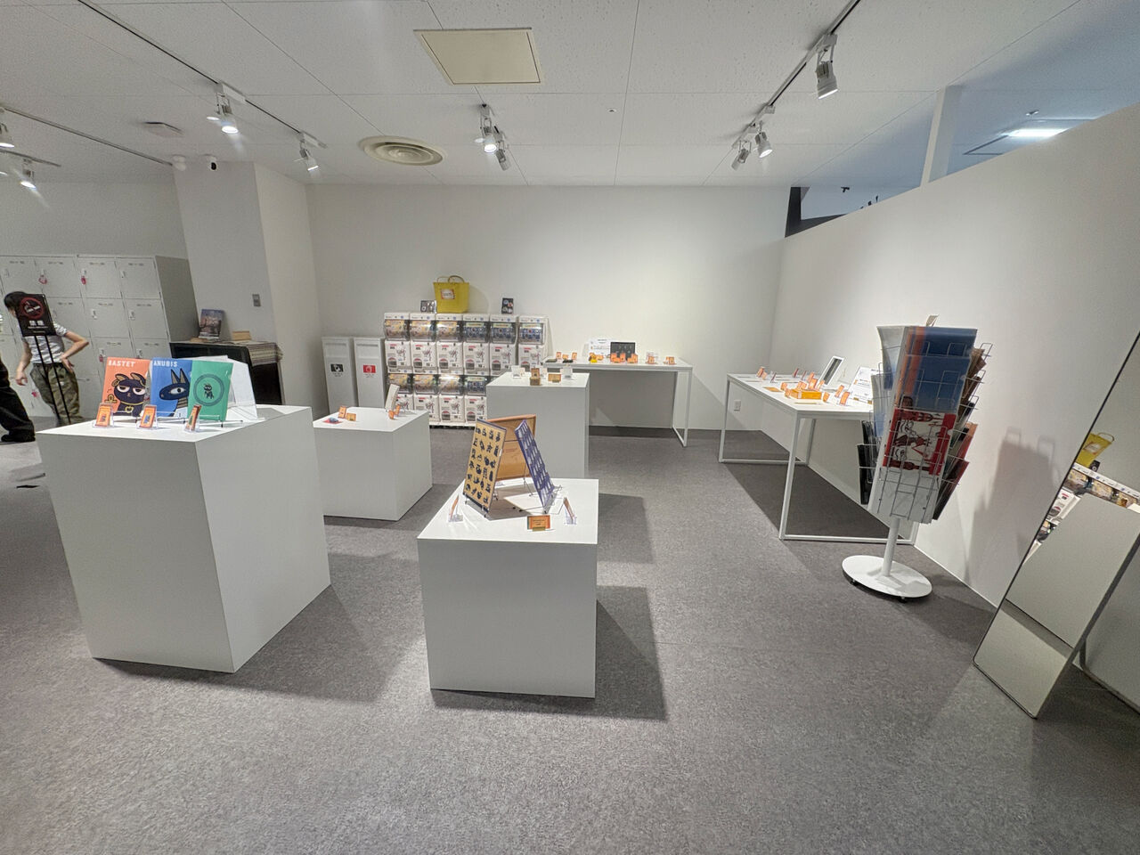 お土産コーナー、薄い灰色のカーペットの上に白い四角い箱上のものとテーブル、テーブルと箱の上には商品が並ぶ、奥の壁沿いにはガチャガチャも
