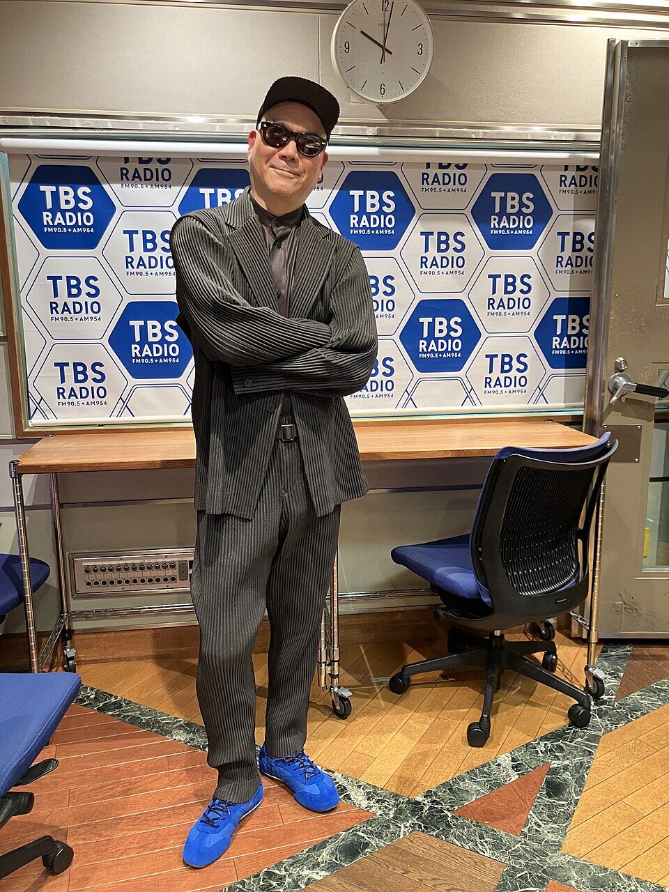 帽子を被ったライムスター宇多丸さんの写真、服は背広、TBSラジオ社内のようで背後のTBSラジオのロゴが沢山書かれた壁がある