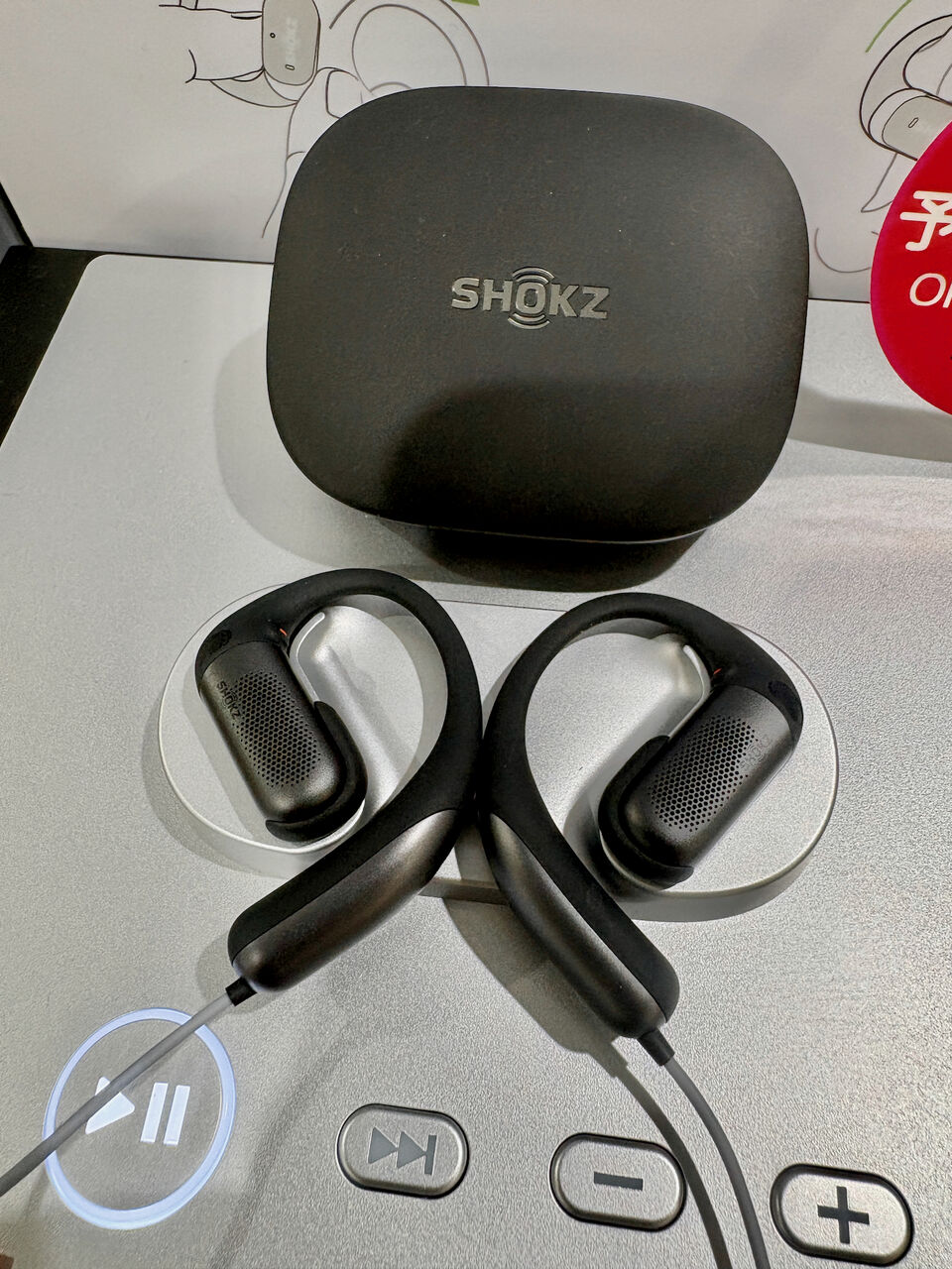 黒色モデル のOpenFit Proの試聴機の写真、ケースの下に左右イヤホンが置いてあり、細いケーブルで繋がれてる、その下に操作ボタン、左端の再生・停止ボタンは光ってる