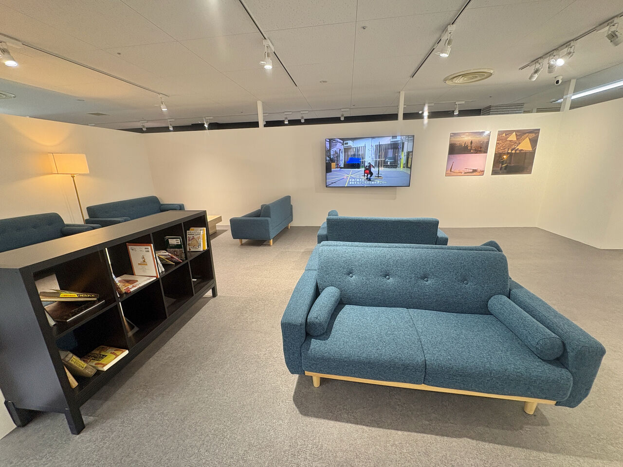 白い壁と薄い灰色のカーペットが敷かれた部屋、右手前から左手奥にかけて複数のソファー、一番手前のソファーの横には黒い低い本棚、本棚には複数の本が置かれてる、奥の壁にはピラミッドのイラストとモニター