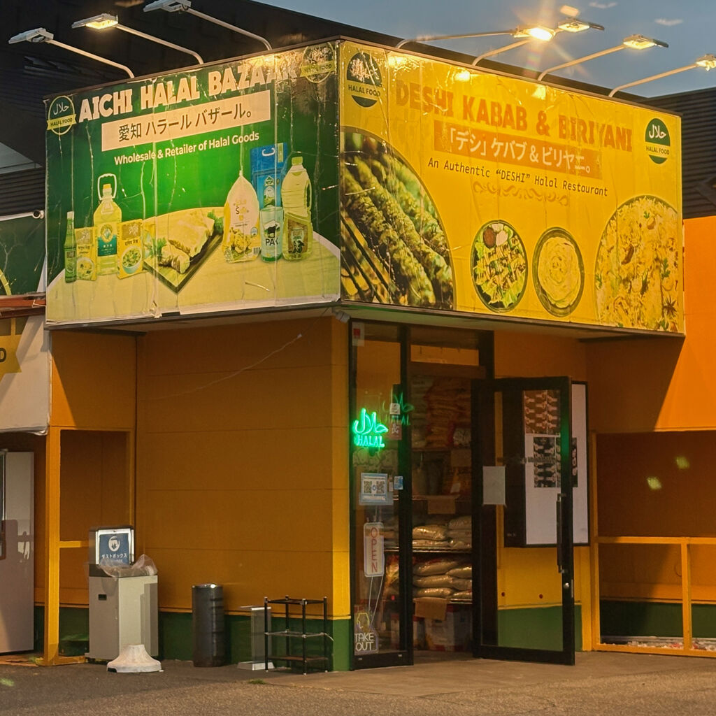 お店の入口アップ写真。入口上に英語と日本語で「デシ」ケバブ＆ビリヤニと言う文字と料理のイラストが描かれてる。入口の中の棚に食材の袋が大小重ねられて置いてある。
