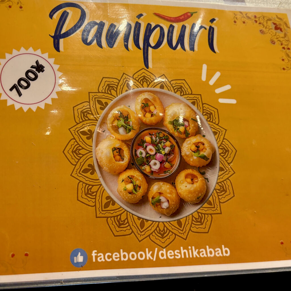 お店に張られてたパニプリのメニュー案内。黄色地で。案内上側に英語で「Panipuri」の文字。その上に唐辛子のイラスト。あと料金700円とFacebookの案内。そして案内中央に植物のような装飾の中央に丸いパニプリの写真。丸い皿の中の周囲に8個の丸いボール状の食べ物。皿の真ん中に野菜や果物を小さく刻んだようなものが小さな丸い皿に入ってる。