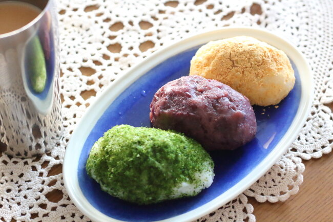 青い楕円形の皿に、緑色の青のりおはぎ、茶色い粒あんおはぎ、黄色のきなこおはぎの3種類が盛り付けられている。手作りおはぎの完成写真
