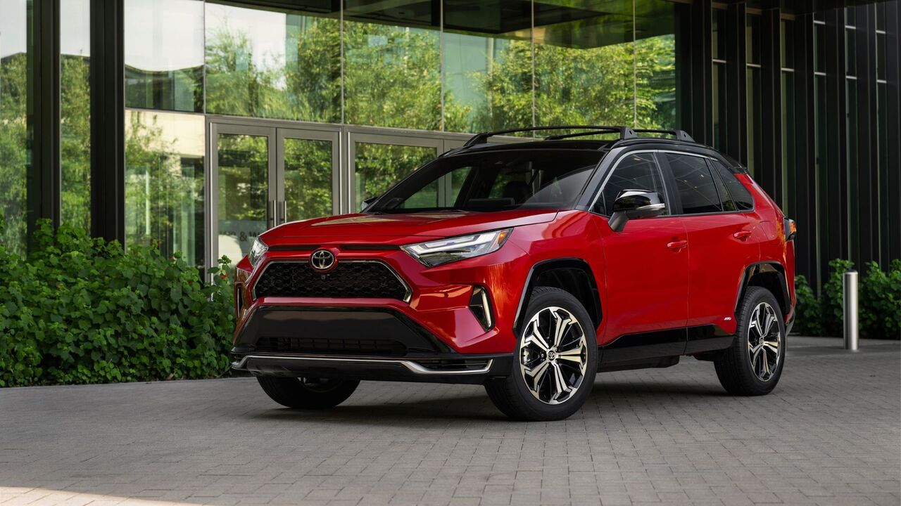 近代的な建物の外に駐車されている赤い 2025 トヨタ RAV4 PHEV XSE の静的なフロント 3/4 ショット。