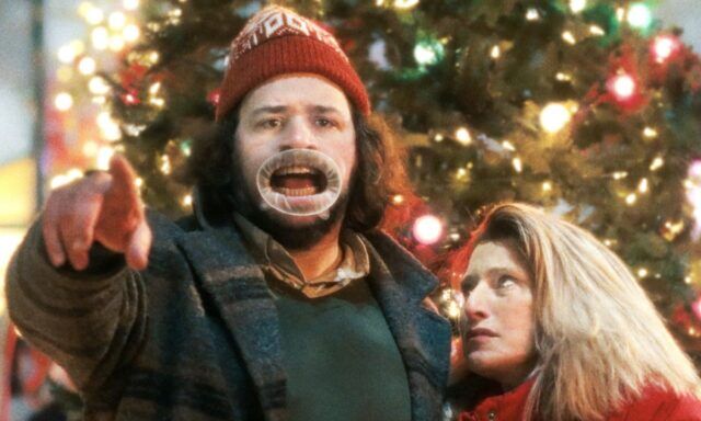 크리스마스 트리 앞에서 어리석은 모자를 쓴 남자가 카메라를 향해 강도질을 하고 있는 동안 여자는 눈을 굴리며 지켜보고 있습니다