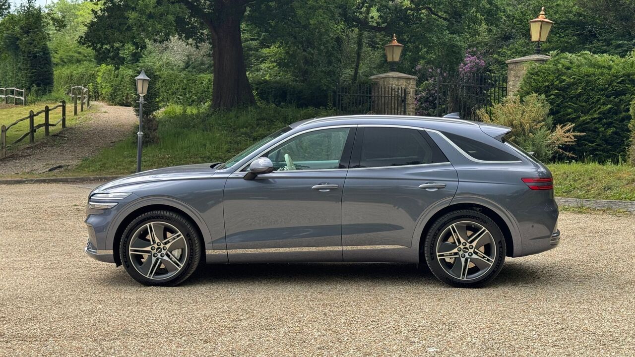 木々を背景に砂利の上に駐車されているグレーの 2026 年型ジェネシス エレクトリファイド GV70 の静的な側面ショット。