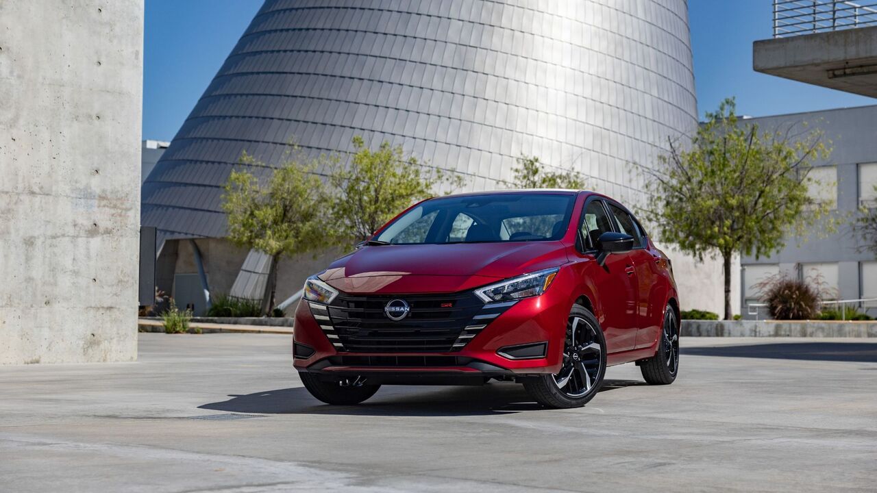 背景にモダンなガラス張りの建物を備えた赤い 2025 Nissan Versa の正面 3/4 ビュー。