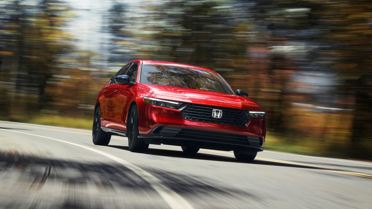 木々が並ぶ田舎道を走る赤い 2024 年型ホンダ アコード スポーツ L のダイナミックなフロントエンドビュー。