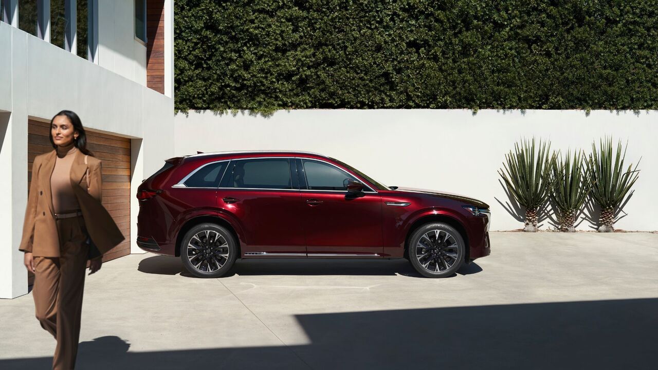 ドライブ中に駐車した赤い 2026 型マツダ CX-90 の静的な横顔ショット。前景を女性が歩いています。