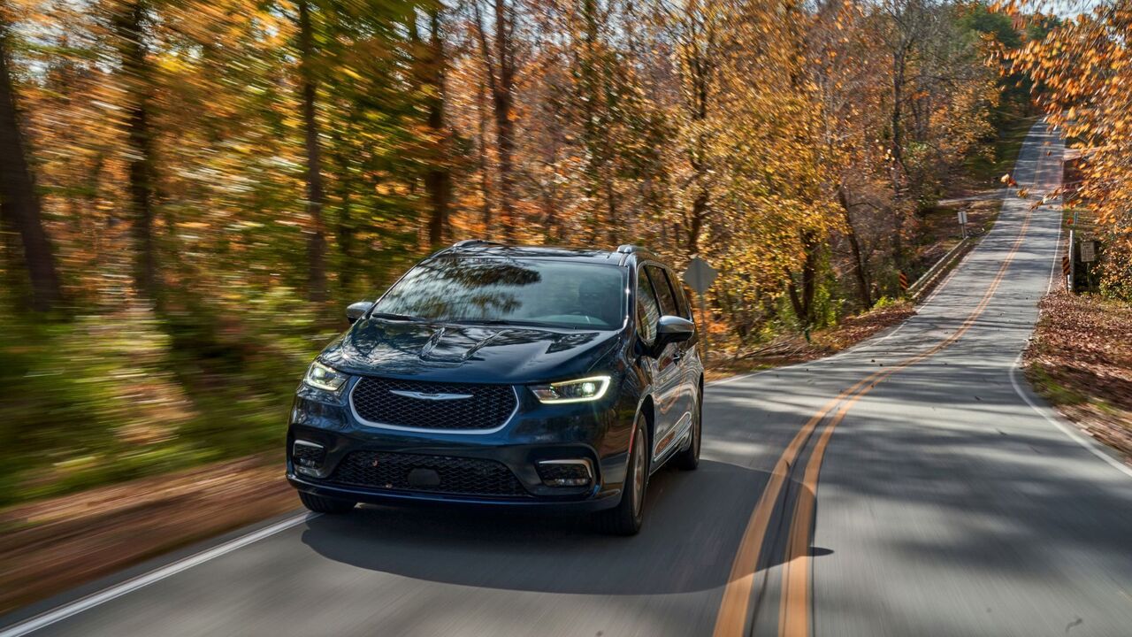 木々が並ぶ田舎道を走行するダークブルーの 2025 年型クライスラー パシフィカ プラグイン ハイブリッド ピナクルのダイナミックなフロント 3/4 ショット。
