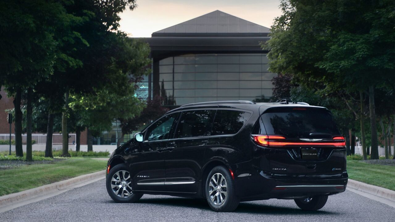モダンな建物と木々を背景にした、黒い 2025 年型クライスラー パシフィカ プラグイン ハイブリッド ピナクルの静止リア 3/4 ショット。