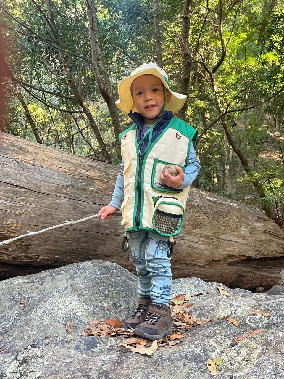 낚시 조끼를 입고 막대기와 바위를 들고 있는 허드슨 오로린(Hudson O'Loughlin)은 쓰러진 통나무와 나무를 배경으로 바위 위에 서 있습니다.